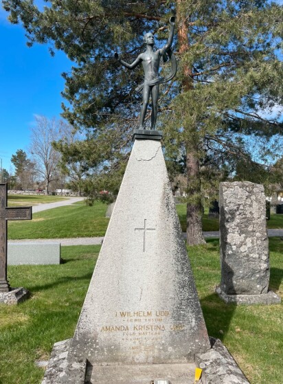 En konformad gravsten med en staty uppe på föreställande en man som ser ut över himlen med höjda händer. Över vänster arm hänger ett tygstycke som vajar i vinden
