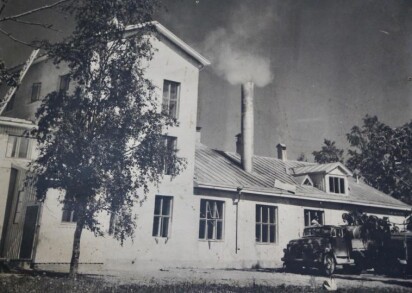 En vit betongbyggnad i envåning med högre del i trevåningar. Det ryker ur en hög rund skorstenspipa.