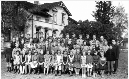 En grupp med barn och några vuxna som poserar sittande och stående framför skolan som är vit.