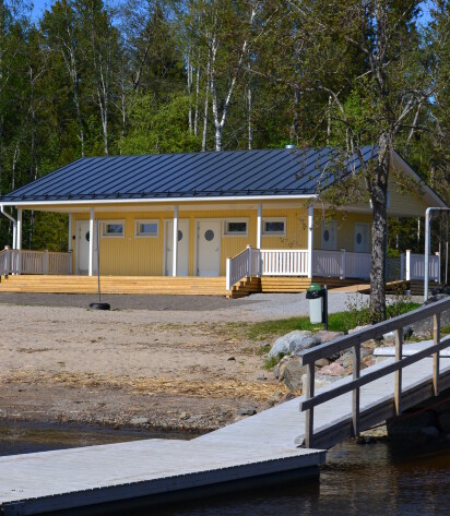 Keltainen pukuhuonerakennus, jossa on valkoinen kaide. Etualalla näkyy laituria ja uimarantaa.