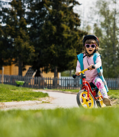 En liten glad flicka klädd i blommiga byxor, prickig rosa tröja och ljusblå reflexväst, solglasögon och röd hjälm cyklar på en liten röd cykel med gula hjul